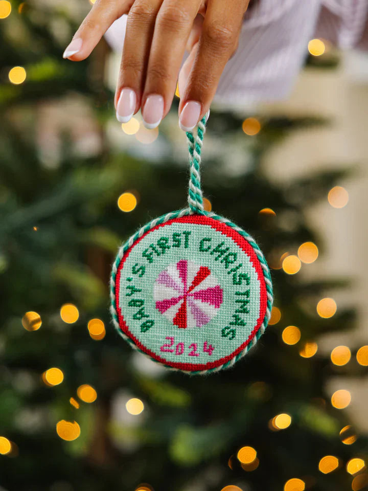 Needlepoint ornament with "Baby's 1st Christmas" text, held in front of a decorated Christmas tree.