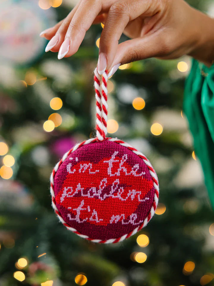 Needlepoint ornament with holiday design, backed in luxury velvet, held in front of festive tree.